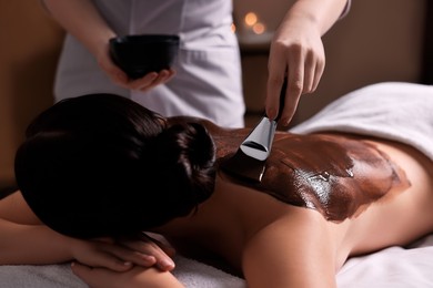 Photo of Chocolate body wrap. Spa worker applying mask onto woman's back in salon, closeup