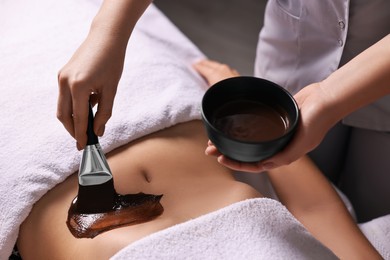 Photo of Chocolate body wrap. Spa worker applying mask onto woman's belly in salon, closeup