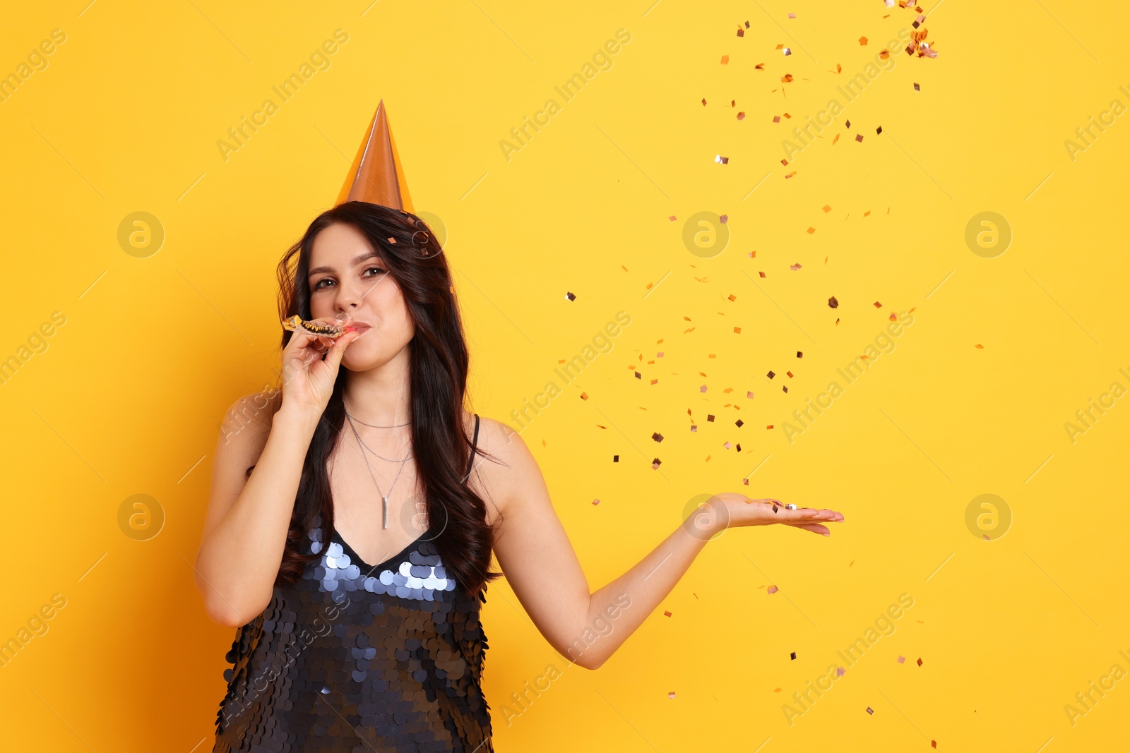 Photo of Woman in conical paper hat with blower and flying confetti on orange background, space for text. Surprise party