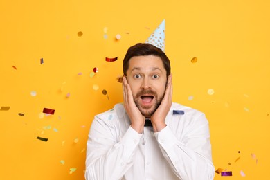 Emotional man in conical paper hat and flying confetti on orange background. Surprise party