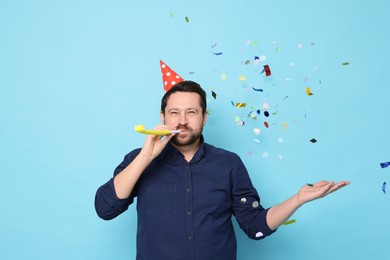 Man in conical paper hat with blower and flying confetti on light blue background. Surprise party