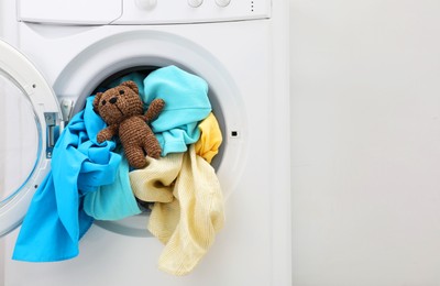 Photo of Washing machine with clothes and toy bear near light wall, space for text