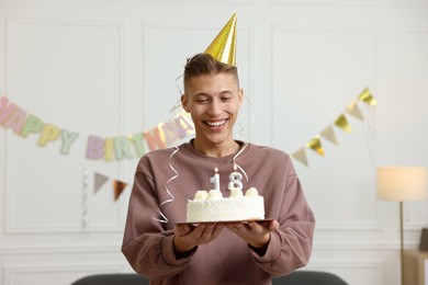 Photo of Coming of age party - 18th birthday. Happy young man in hat holding tasty cake with number shaped candles at home