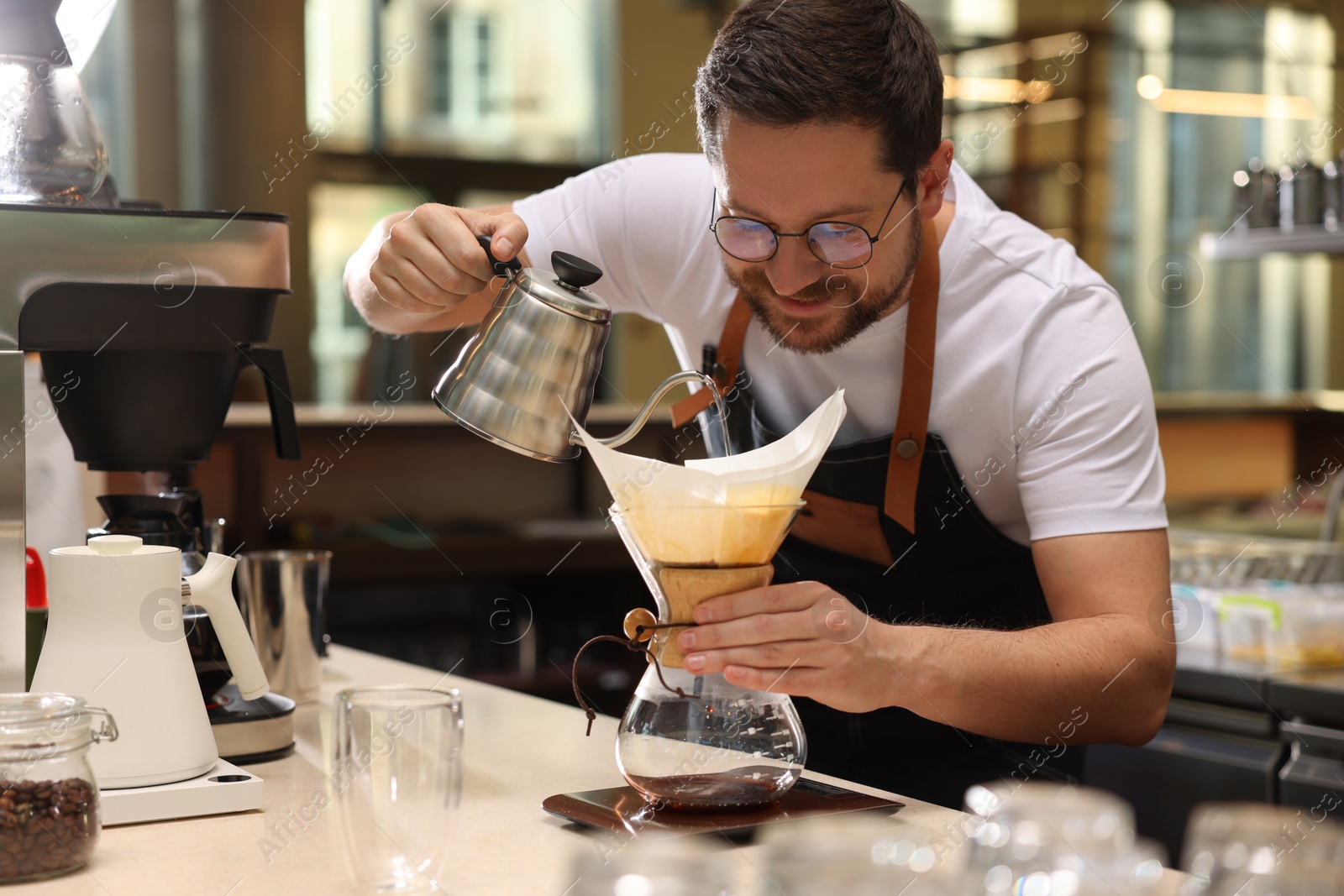 Photo of Barista brewing coffee in glass coffeemaker with paper filter at table in cafe