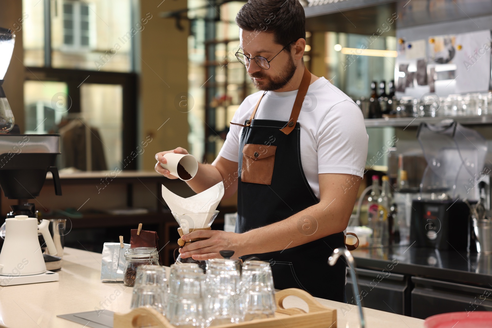 Photo of Barista pouring ground coffee into glass coffeemaker with paper filter at table in cafe