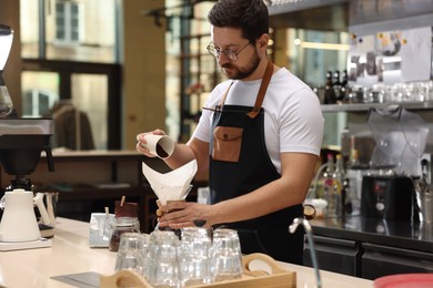 Photo of Barista pouring ground coffee into glass coffeemaker with paper filter at table in cafe