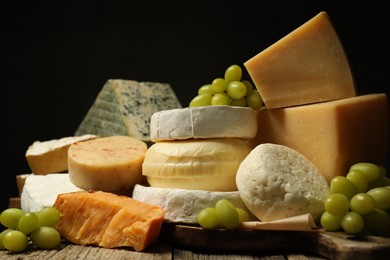 Photo of Different types of cheese and grapes on wooden table against black background, closeup