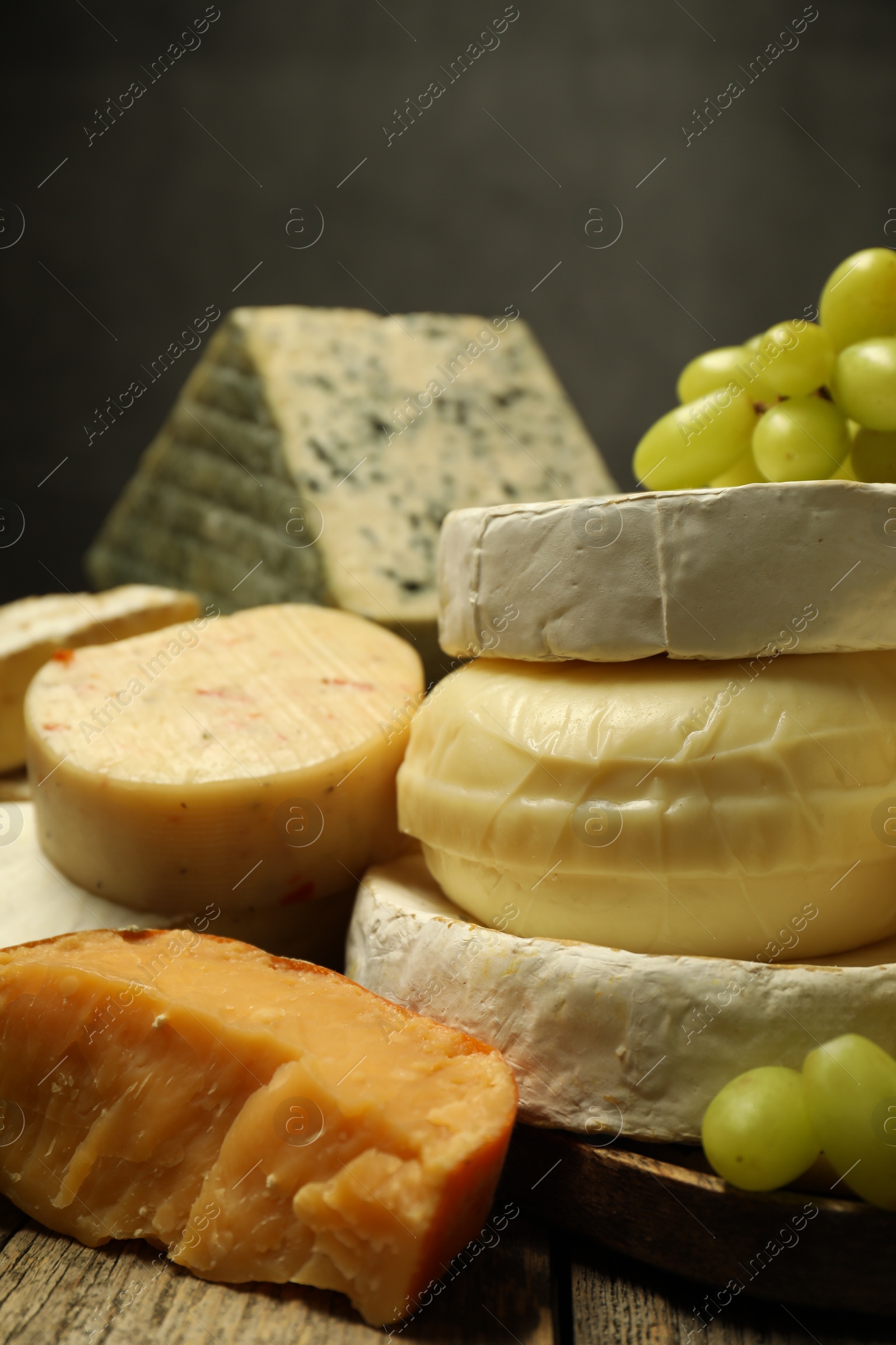 Photo of Different types of cheese and grapes on wooden table against dark background, closeup