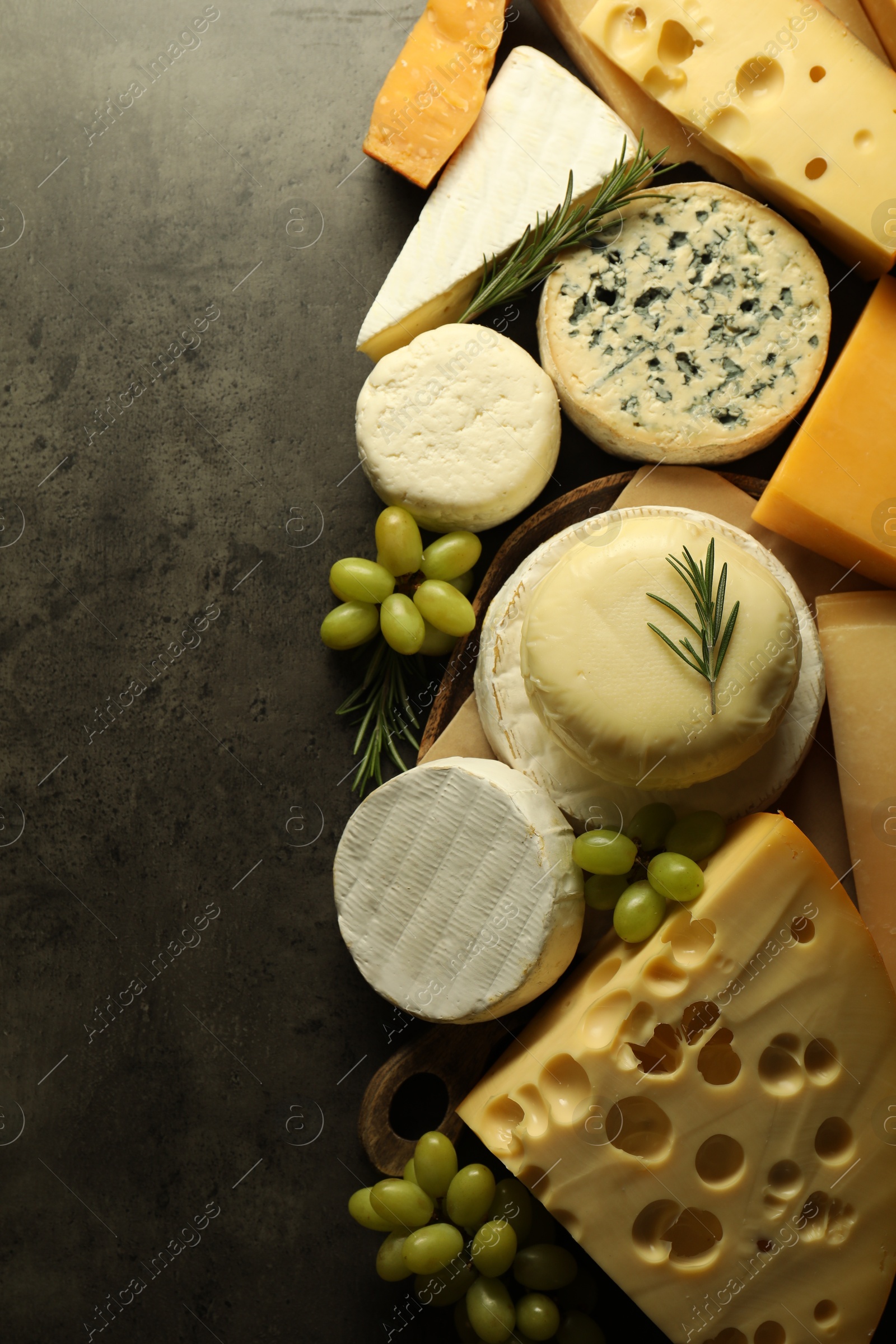 Photo of Different types of cheese, rosemary and grapes on dark textured table, flat lay. Space for text