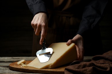 Photo of Woman cutting delicious cheese with slicer at wooden table against black background, closeup