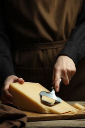 Photo of Woman cutting delicious cheese with slicer at wooden table against black background, closeup