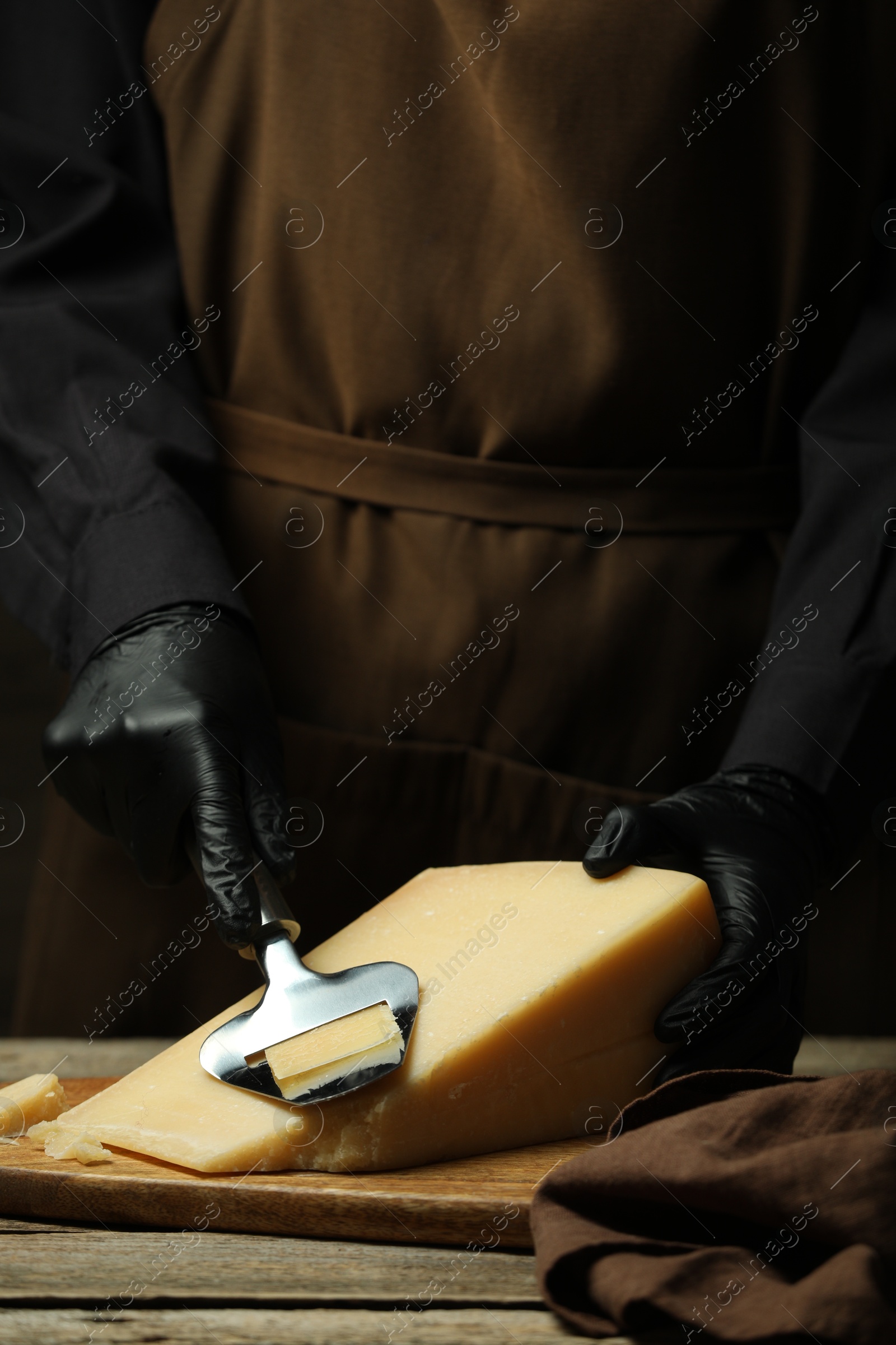 Photo of Woman in gloves cutting delicious cheese with slicer at wooden table, closeup