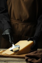 Photo of Woman in gloves cutting delicious cheese with slicer at wooden table, closeup