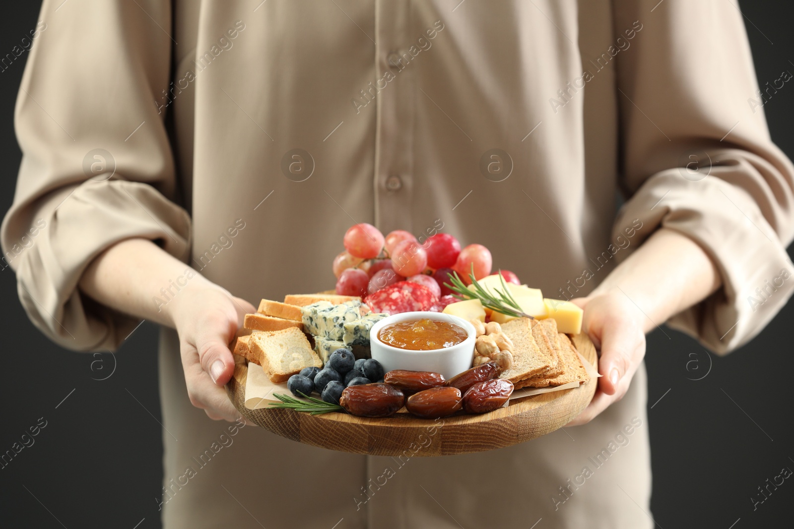 Photo of Woman holding board with different types of delicious cheese and other snacks on grey background, closeup