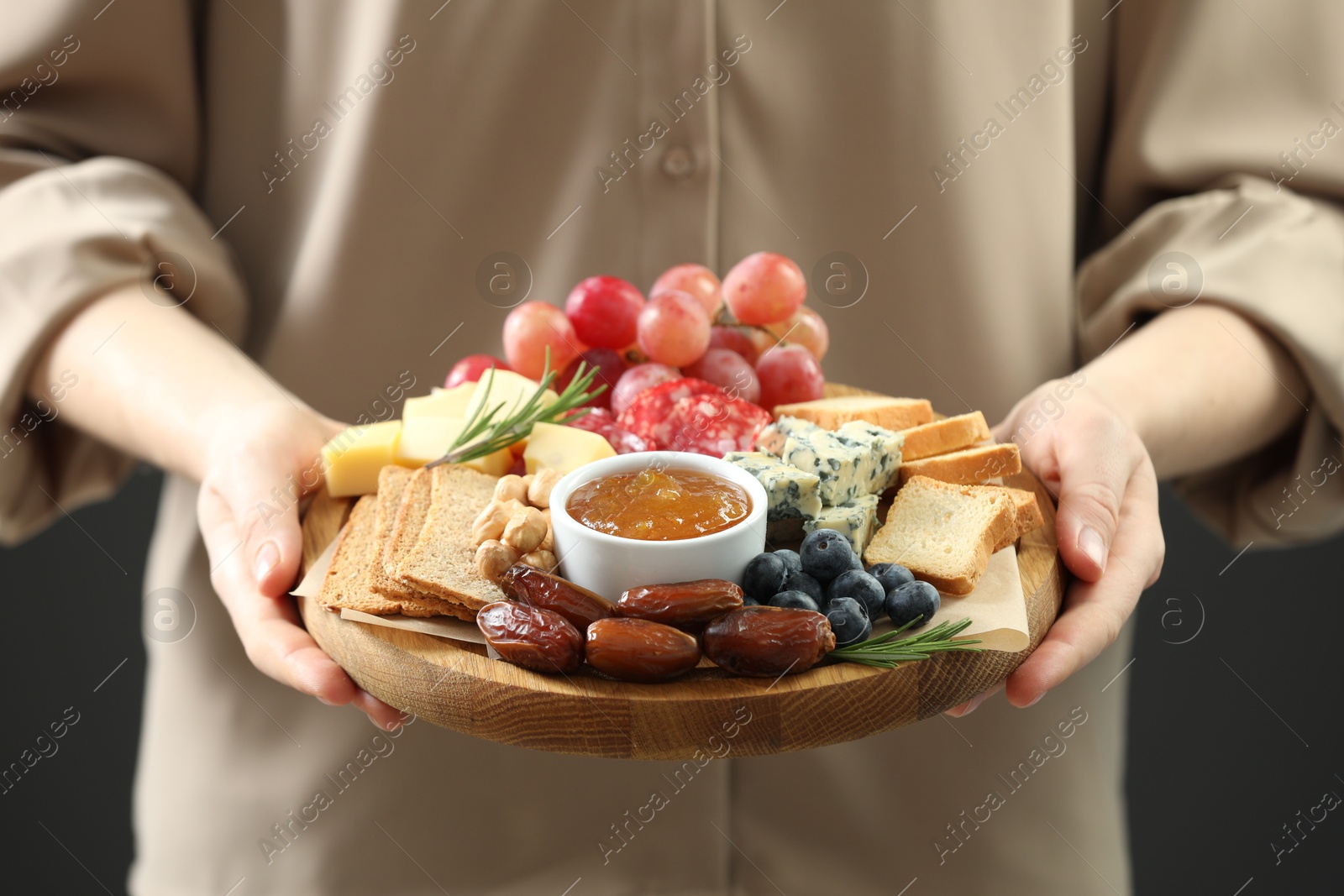 Photo of Woman holding board with different types of delicious cheese and other snacks on grey background, closeup