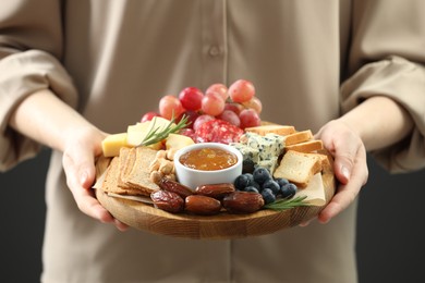 Photo of Woman holding board with different types of delicious cheese and other snacks on grey background, closeup