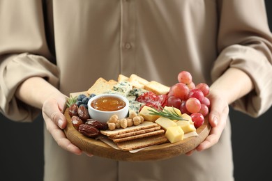 Photo of Woman holding board with different types of delicious cheese and other snacks on grey background, closeup