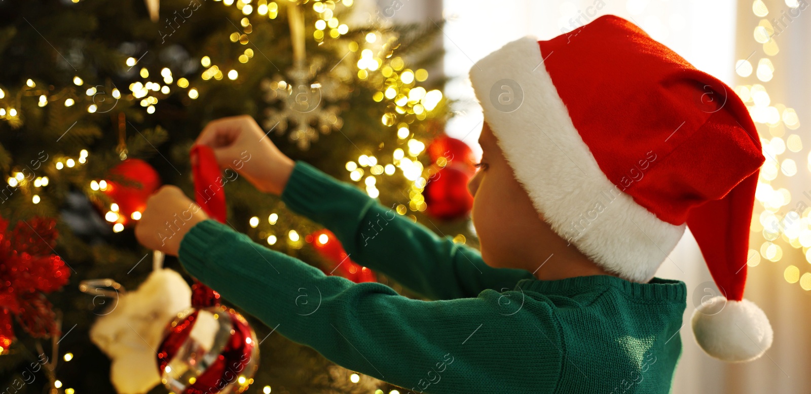 Photo of Cute little boy in Santa hat decorating Christmas tree at home