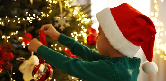 Photo of Cute little boy in Santa hat decorating Christmas tree at home