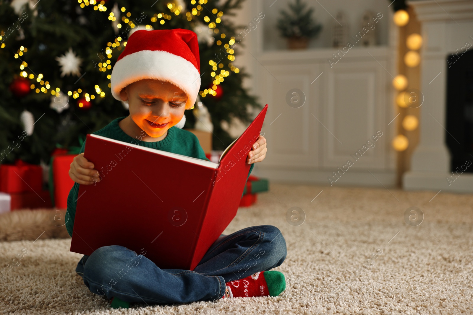 Photo of Cute little boy with Santa hat reading book on rug in room decorated for Christmas, space for text