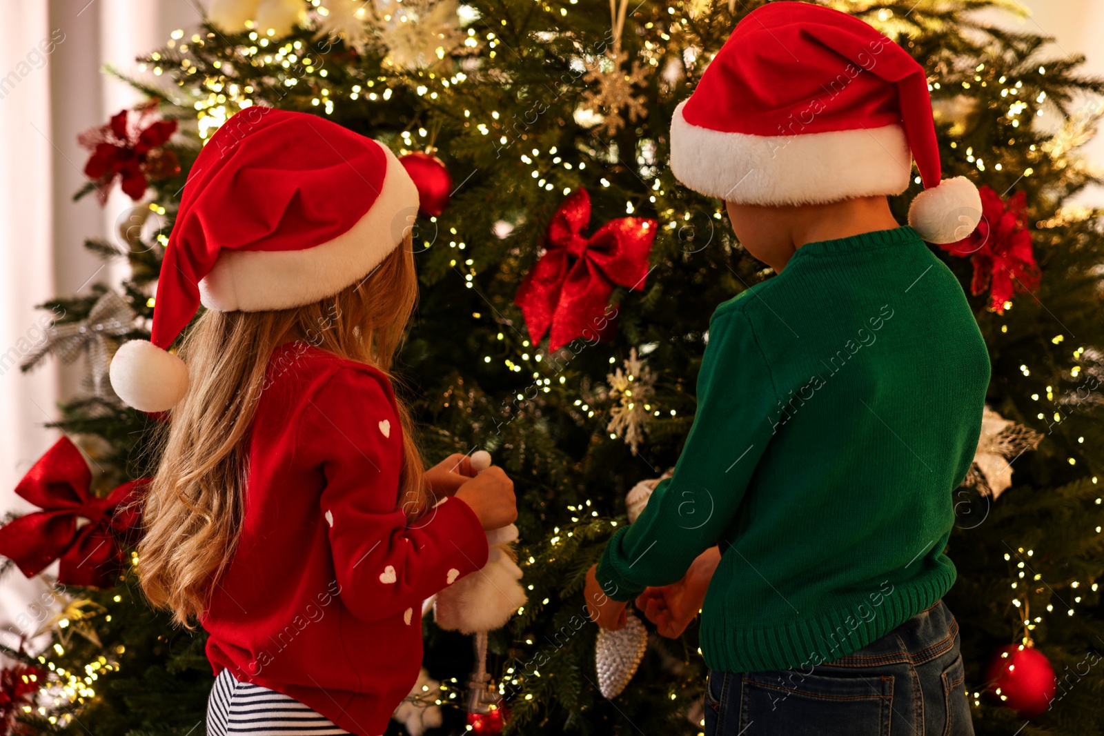 Photo of Little kids in Santa hats decorating Christmas tree at home