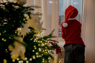 Photo of Little boy with Santa hat near window in room decorated for Christmas, back view