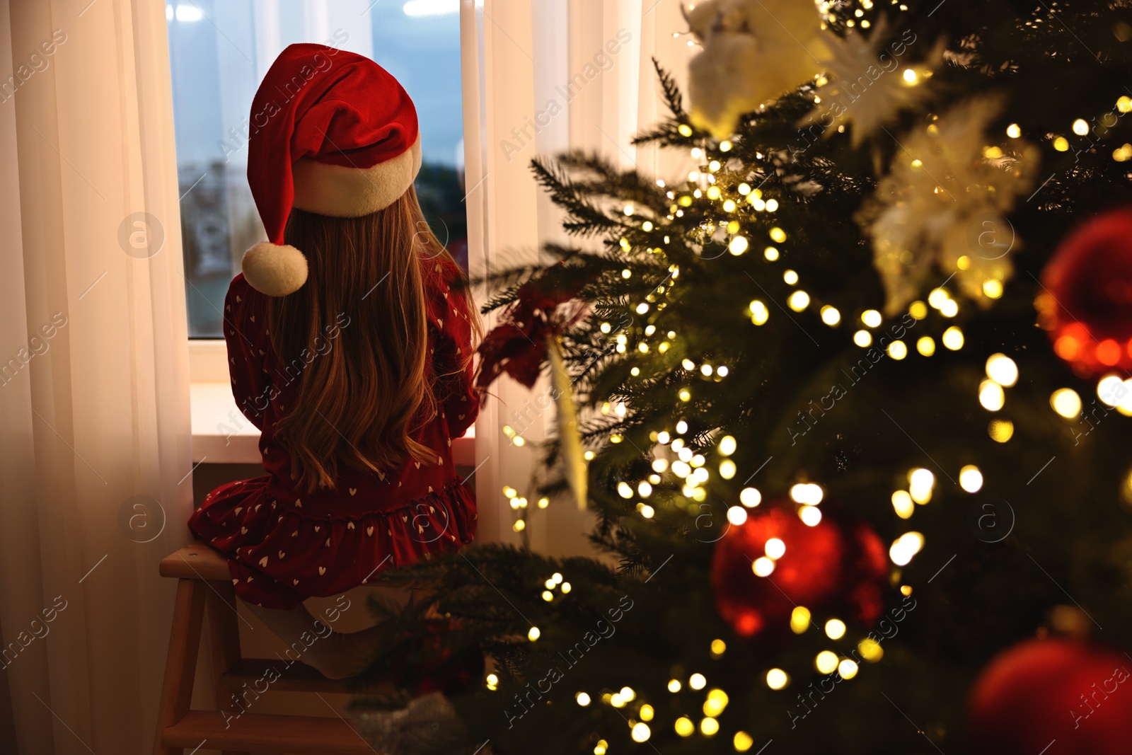 Photo of Little girl with Santa hat near window in room decorated for Christmas, back view