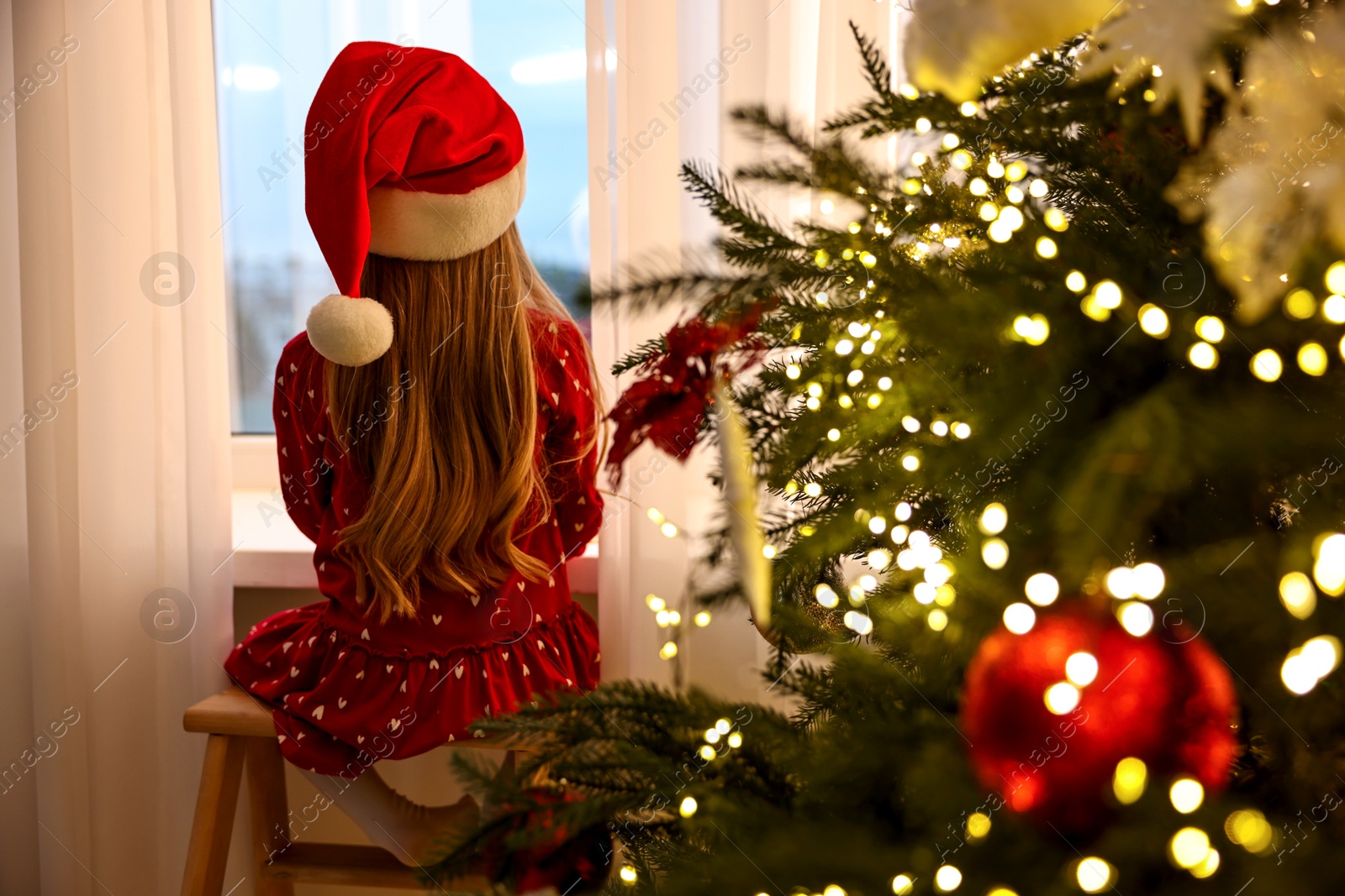 Photo of Little girl with Santa hat near window in room decorated for Christmas, back view