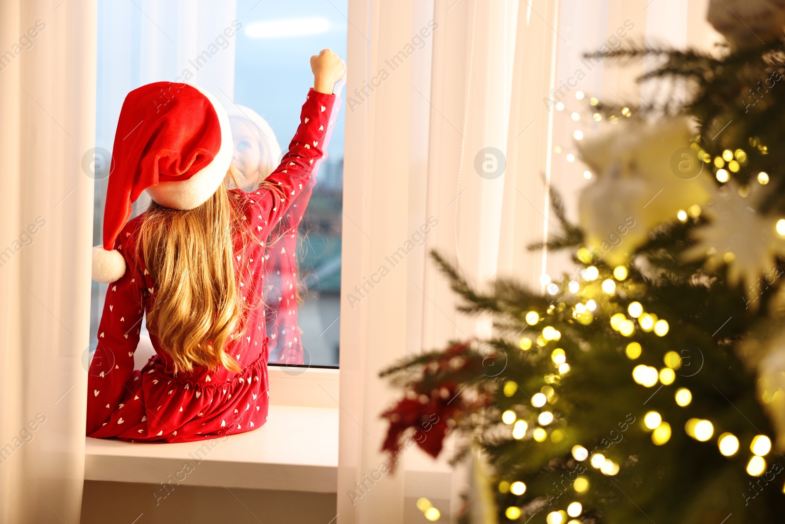 Photo of Little girl with Santa hat near window in room decorated for Christmas, back view