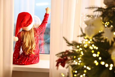 Photo of Little girl with Santa hat near window in room decorated for Christmas, back view