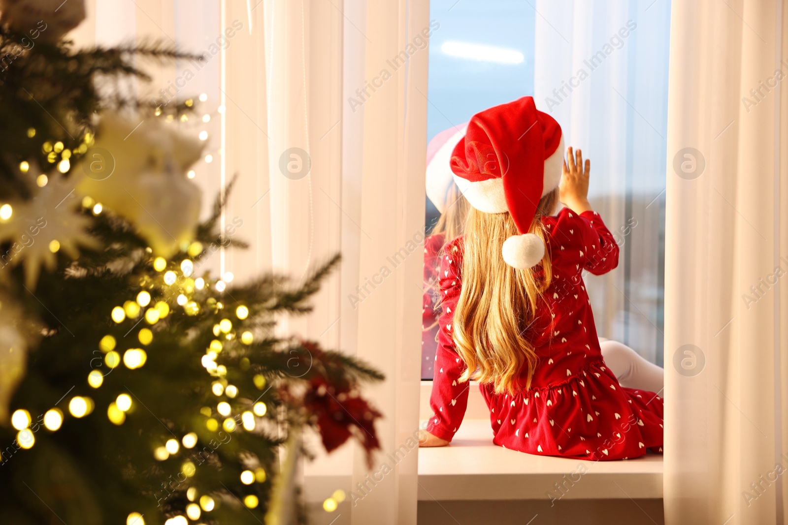 Photo of Little girl with Santa hat near window in room decorated for Christmas, back view