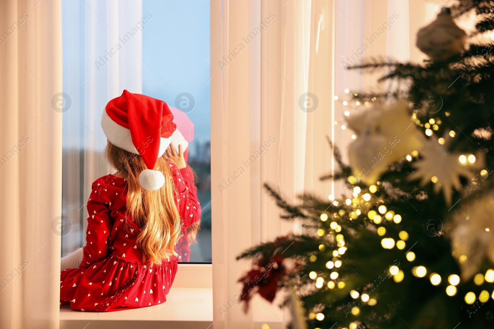 Photo of Little girl with Santa hat near window in room decorated for Christmas, back view