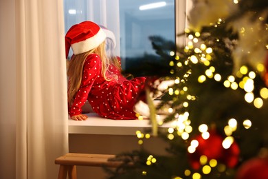 Photo of Little girl with Santa hat near window in room decorated for Christmas