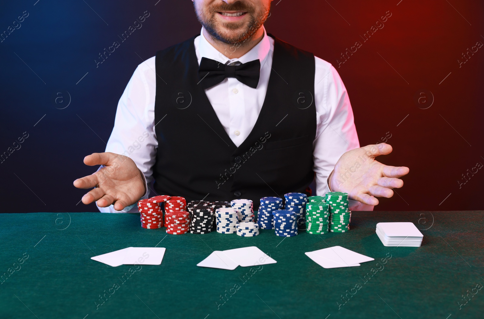 Photo of Professional croupier at table with playing cards and casino chips against color background, closeup