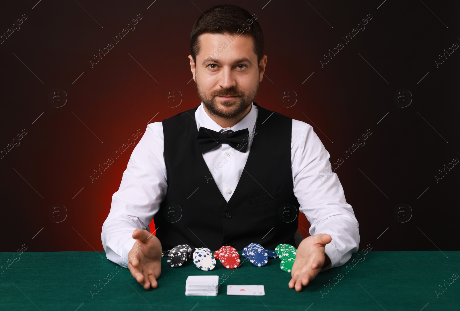 Photo of Professional croupier at gambling table with playing cards and casino chips against dark red background