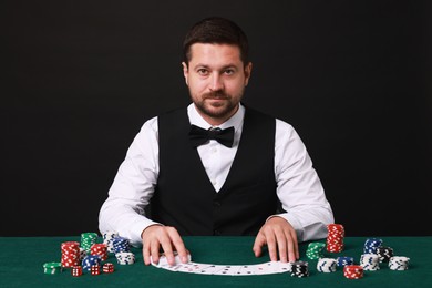 Professional croupier with playing cards at gambling table against black background