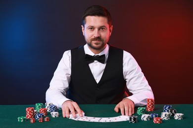 Professional croupier with playing cards at gambling table against color background