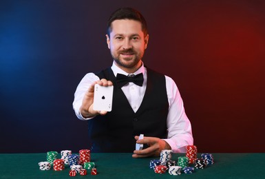 Professional croupier with playing cards at gambling table against color background