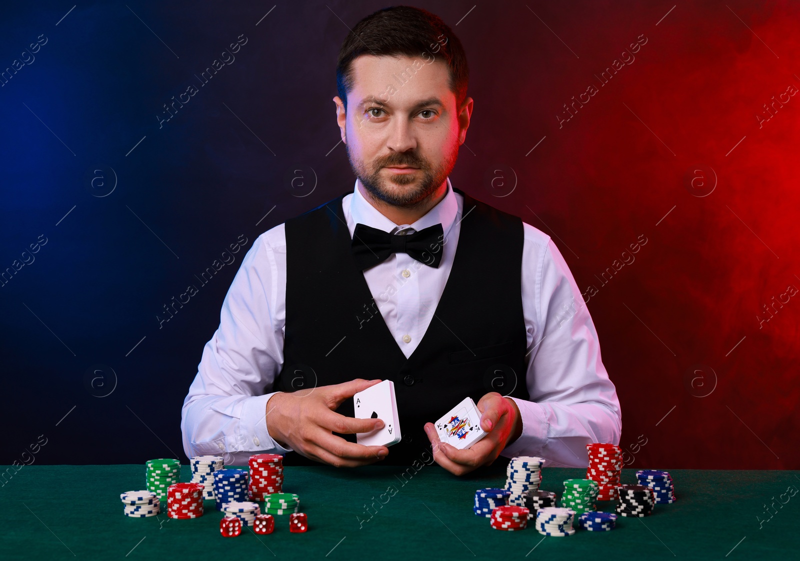 Photo of Professional croupier with playing cards at gambling table against color background with smoke