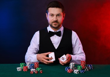 Photo of Professional croupier with playing cards at gambling table against color background with smoke