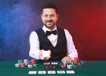 Professional croupier with playing cards at gambling table against color background with smoke