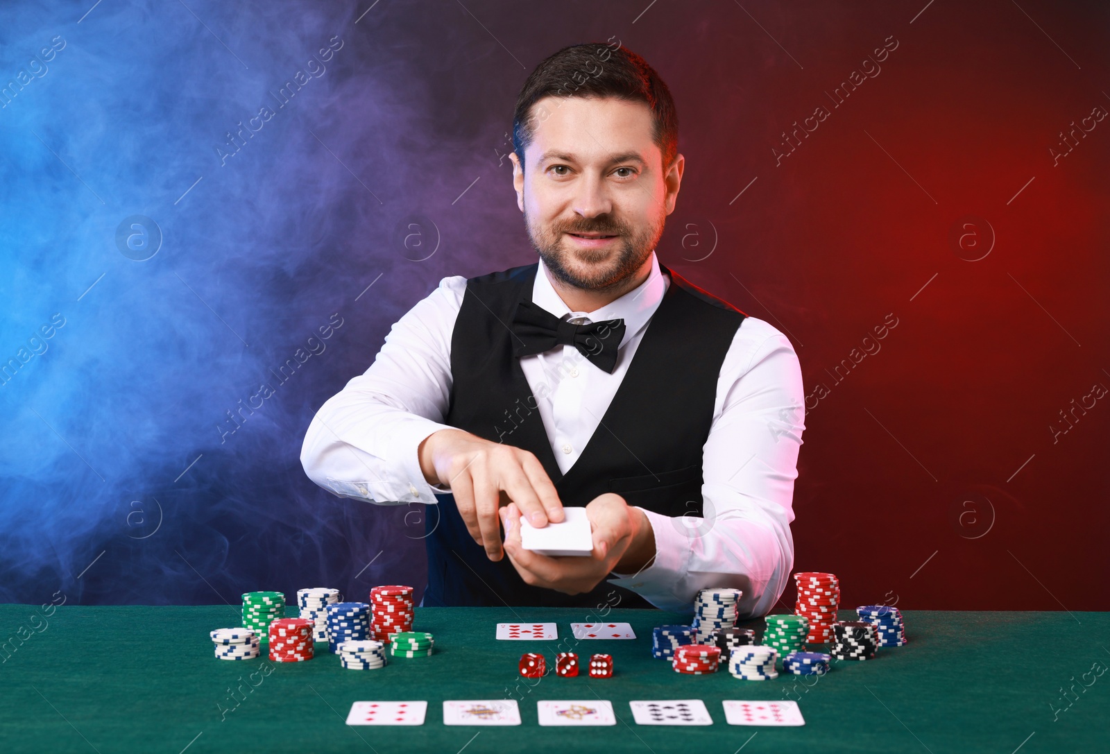 Photo of Professional croupier with playing cards at gambling table against color background with smoke