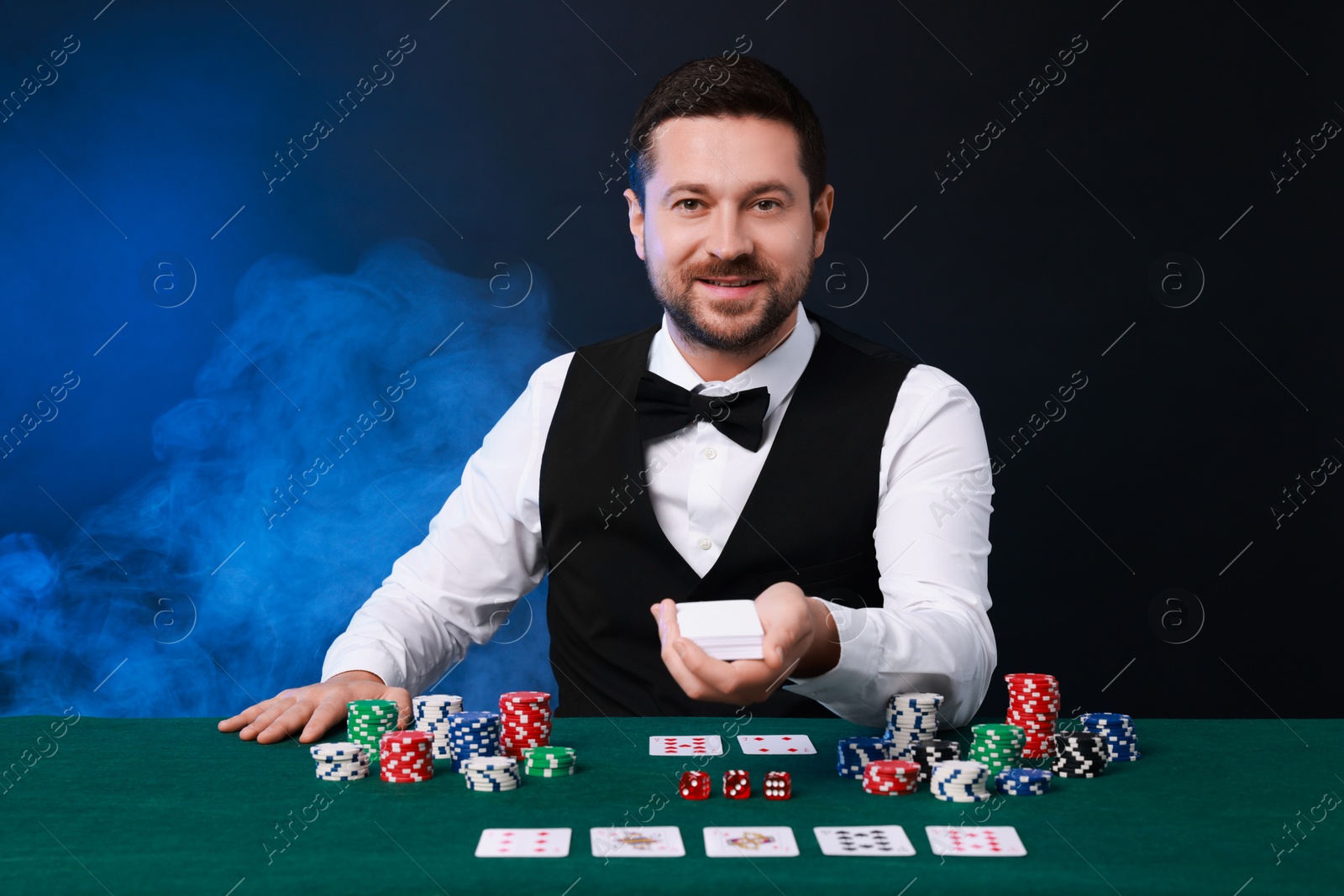 Photo of Professional croupier with playing cards at gambling table against color background with smoke