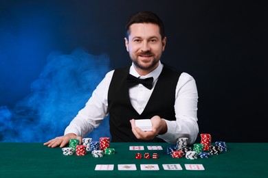 Professional croupier with playing cards at gambling table against color background with smoke