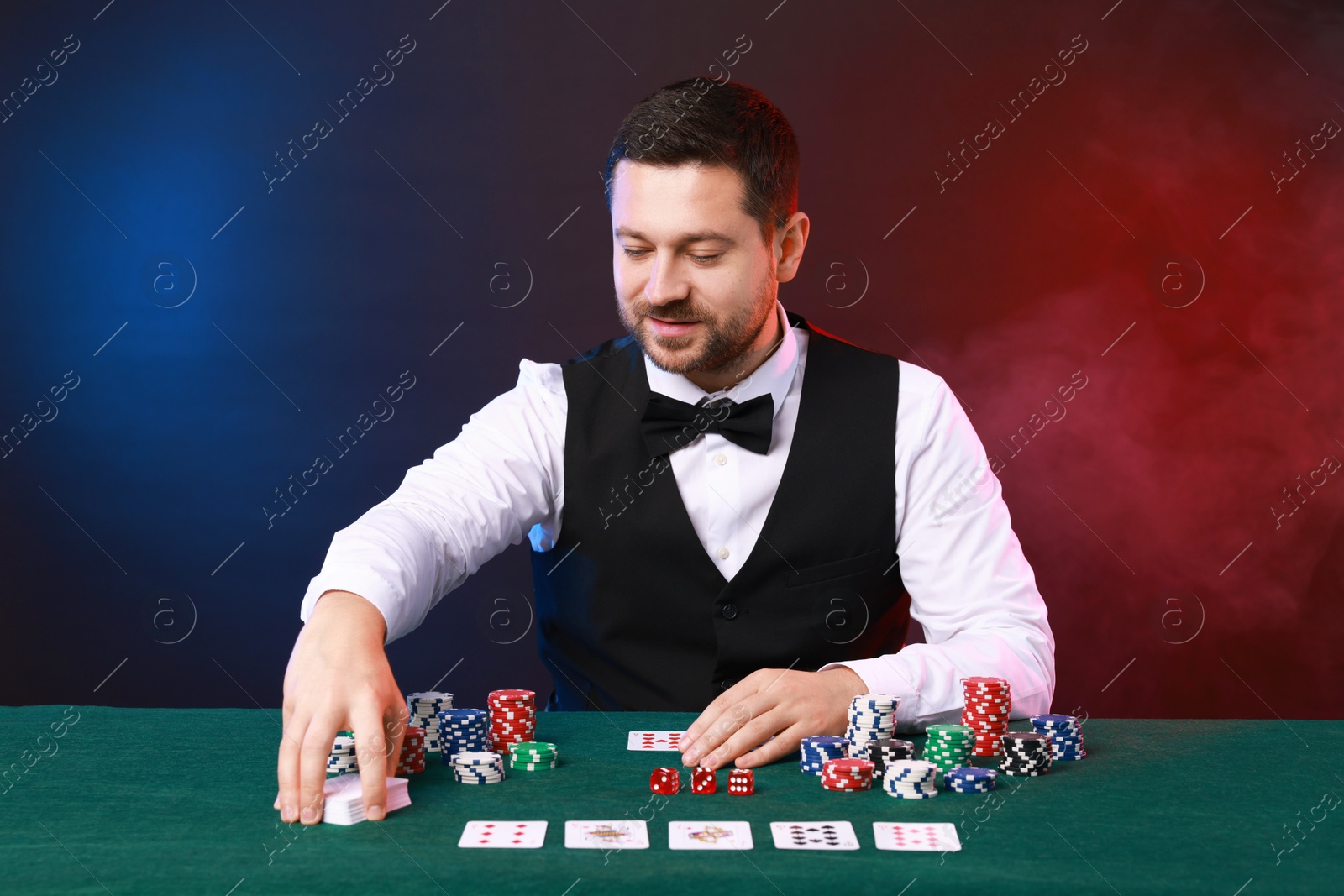 Photo of Professional croupier at gambling table with playing cards, casino chips and dice against color background with smoke