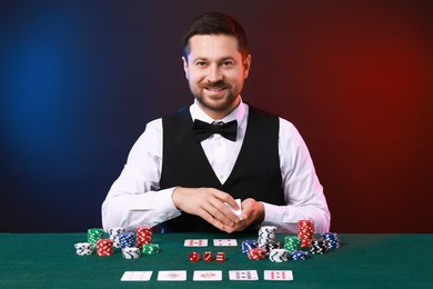 Professional croupier with playing cards at gambling table against color background