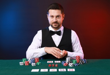 Professional croupier with playing cards at gambling table against color background