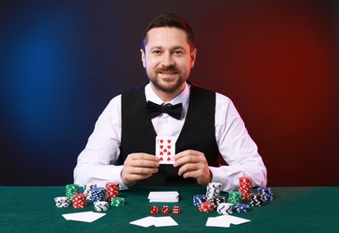 Professional croupier with playing card at gambling table against color background