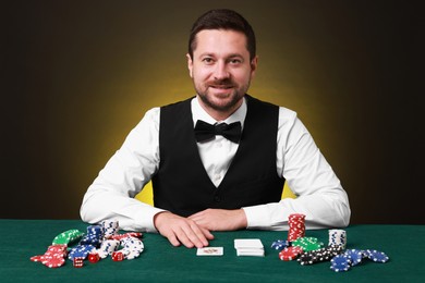 Photo of Professional croupier at gambling table with playing cards, casino chips and dice against dark yellow background