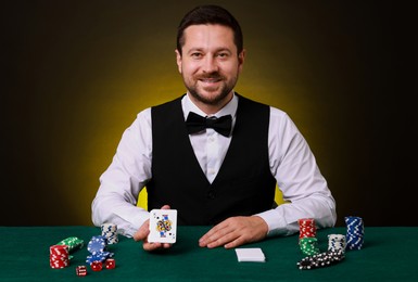 Professional croupier with playing cards at gambling table on dark yellow background